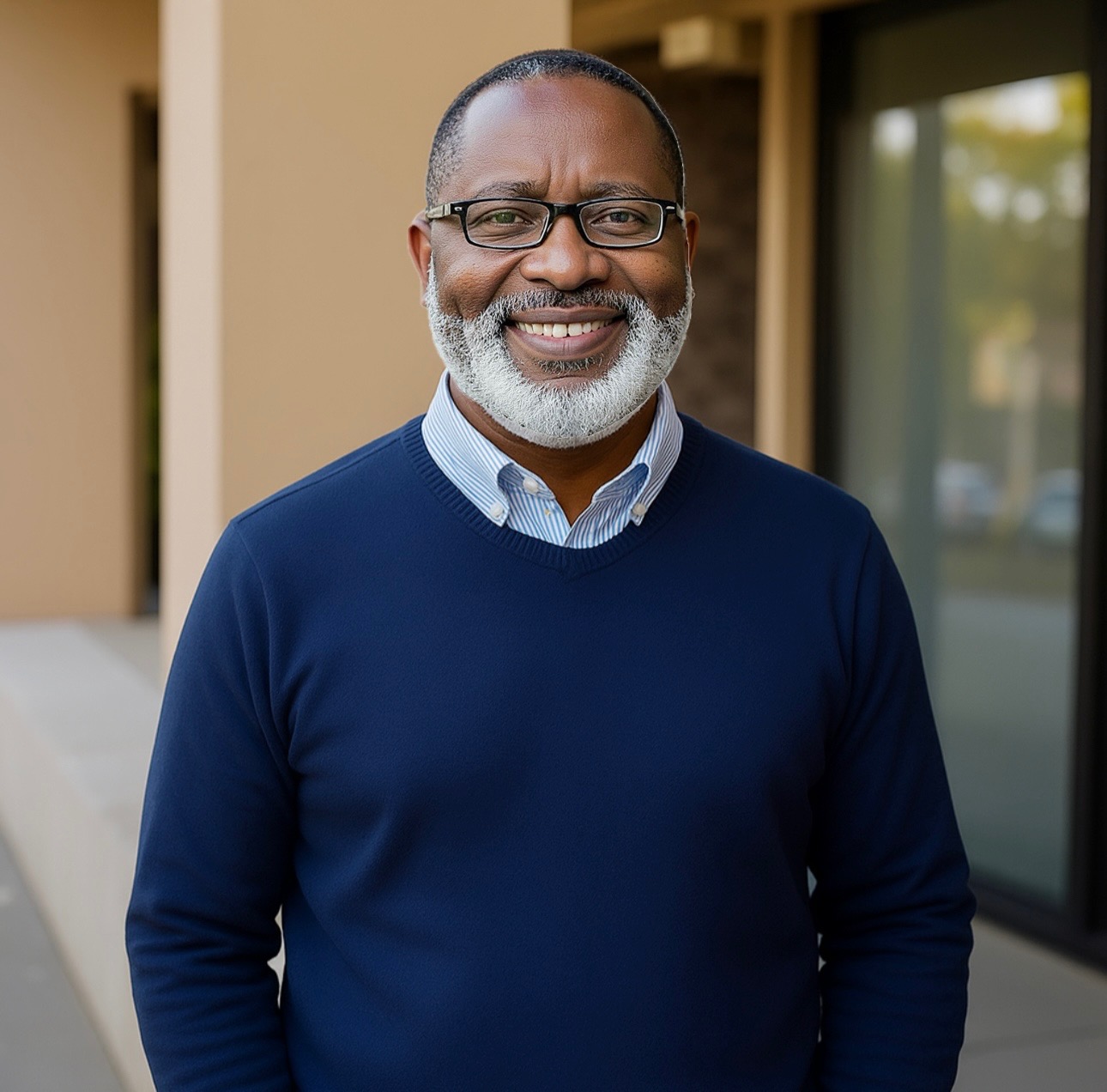 Smiling man with a beard wearing glasses and a navy sweater, standing outdoors in front of a building.
