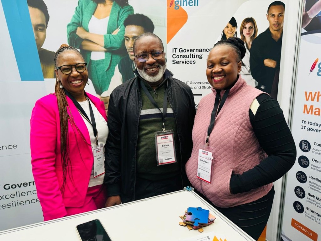Three professionals posing for a photo at an event, smiling and wearing name tags. The background features a poster for IT governance consulting services.